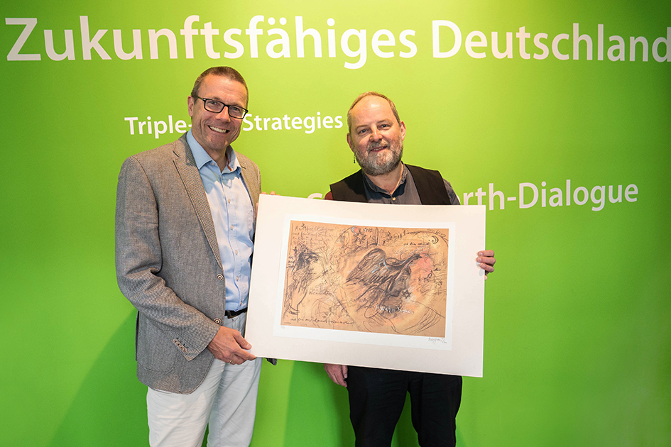 Prof. Dr. Uwe Schneidewind, Präsident des Wuppertal Instituts, und der Komponist Ulrich Klan, der die musikalische Leitung des Zukunftsfestivals übernimmt, zeigen einen Kunstdruck der Künstlerin Ulle Hees aus dem Jahr 2000.